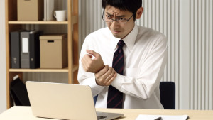 DALL·E 2024-04-21 23.41.54 - A Japanese businessman experiencing wrist pain while using a computer. He is sitting at a desk in an office environment, looking concerned as he holds